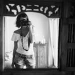 Black and white of casual female photographer in summer clothes standing against mirror in light room and taking photo with camera